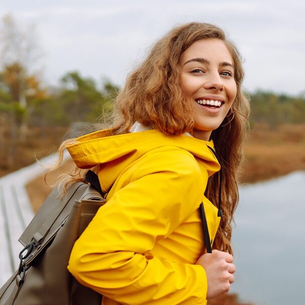 Smiling female traveler in yellow coat walks along wooden path among wild nature. Travel, vacation.