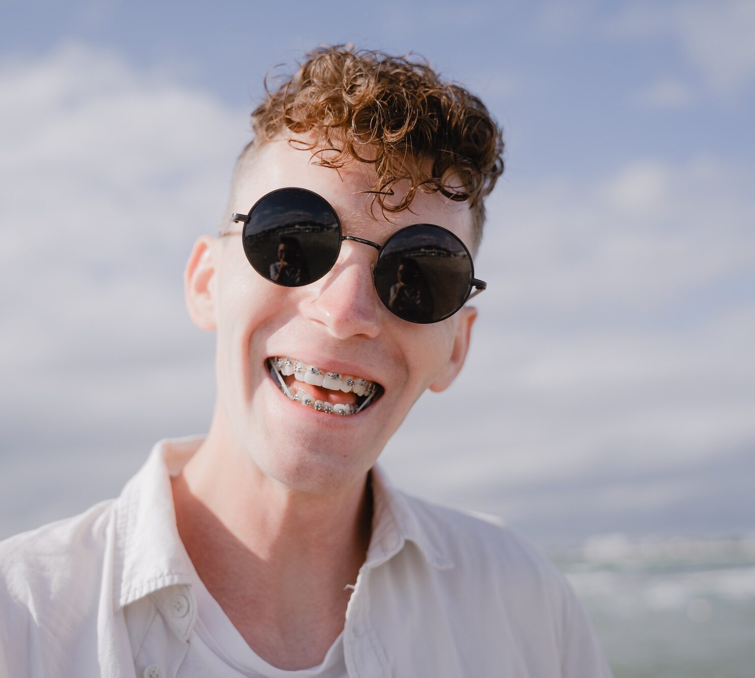 young guy smiling with braces on his teeth, with curly hair and sunglasses